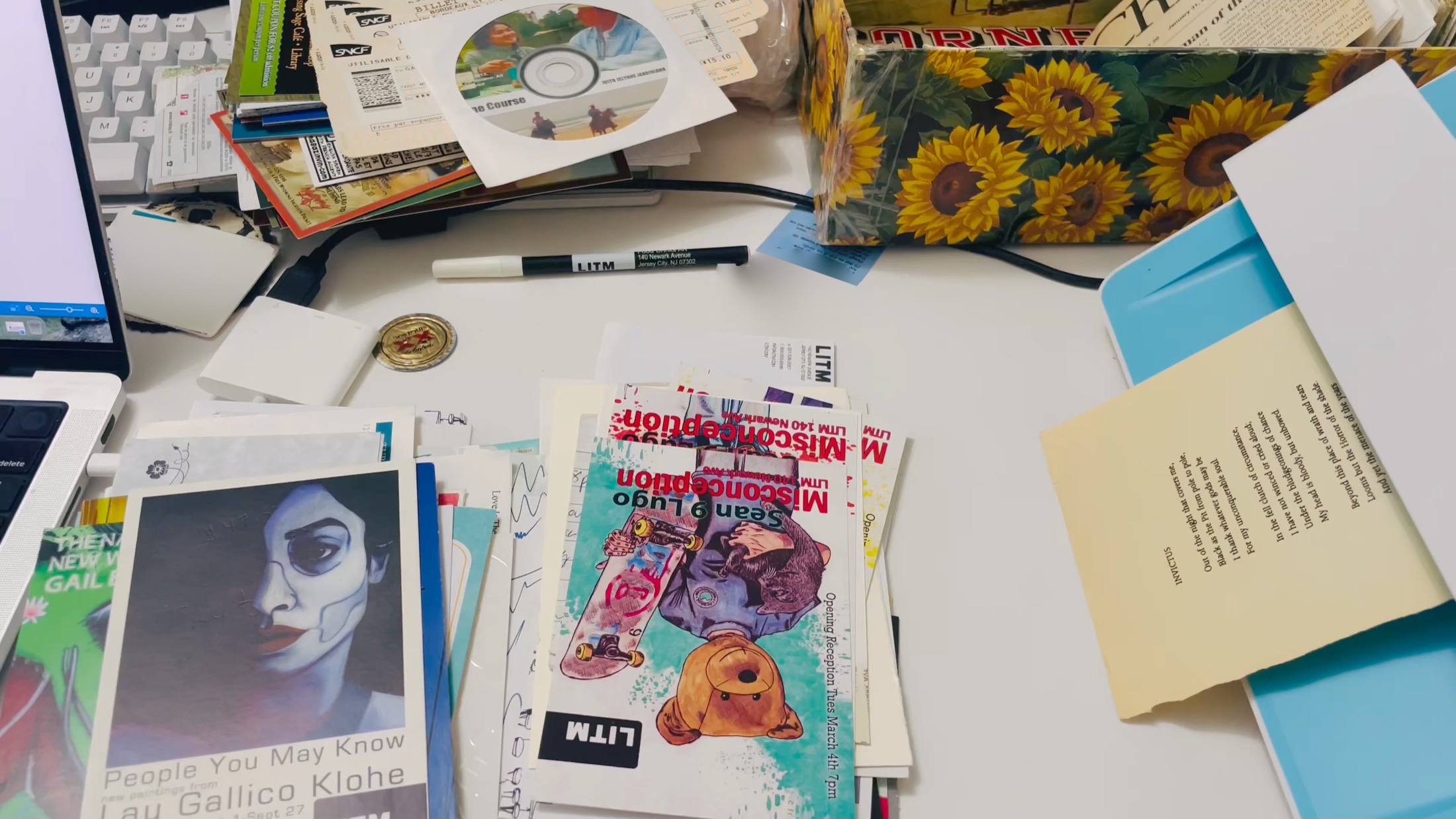 A desk spread with zines, postcards, prints and a printed poem; the sunflower box sits at the far edge.