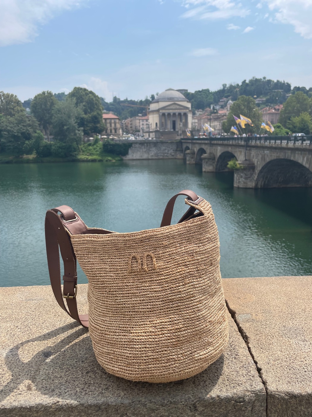 The finished raffia bucket bag with a brown leather strap, resting on a stone ledge above a river.