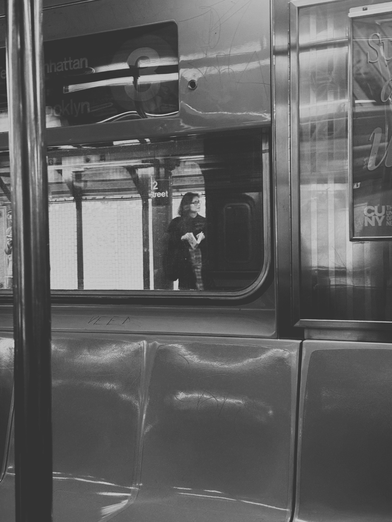Black and white: through the window of a New York subway car, Jelynne stands on the platform, beyond a steel pole and a row of seats.
