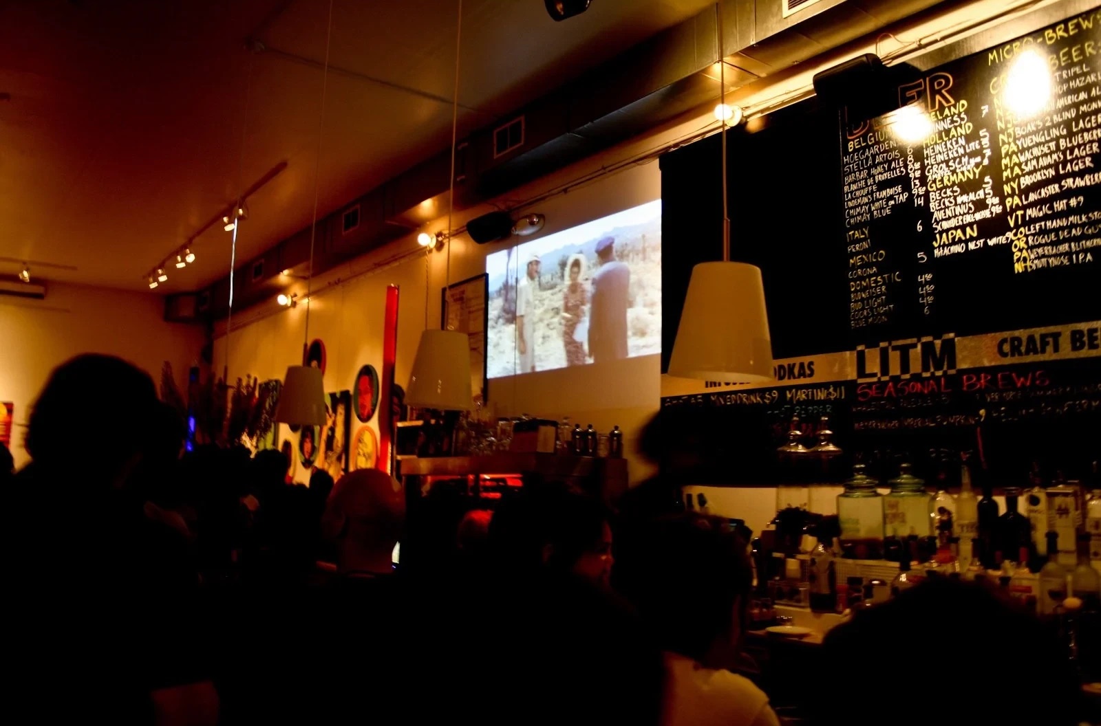 The finished room at night: a crowd in silhouette watches a film projected on the wall, beside a chalkboard beer menu and the LITM sign over the bar.